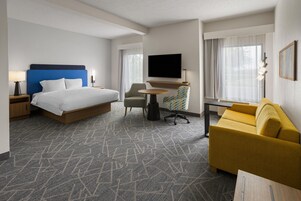 Habitación de hotel con cama, sofá, escritorio, silla, televisión y ventana con cortinas.
