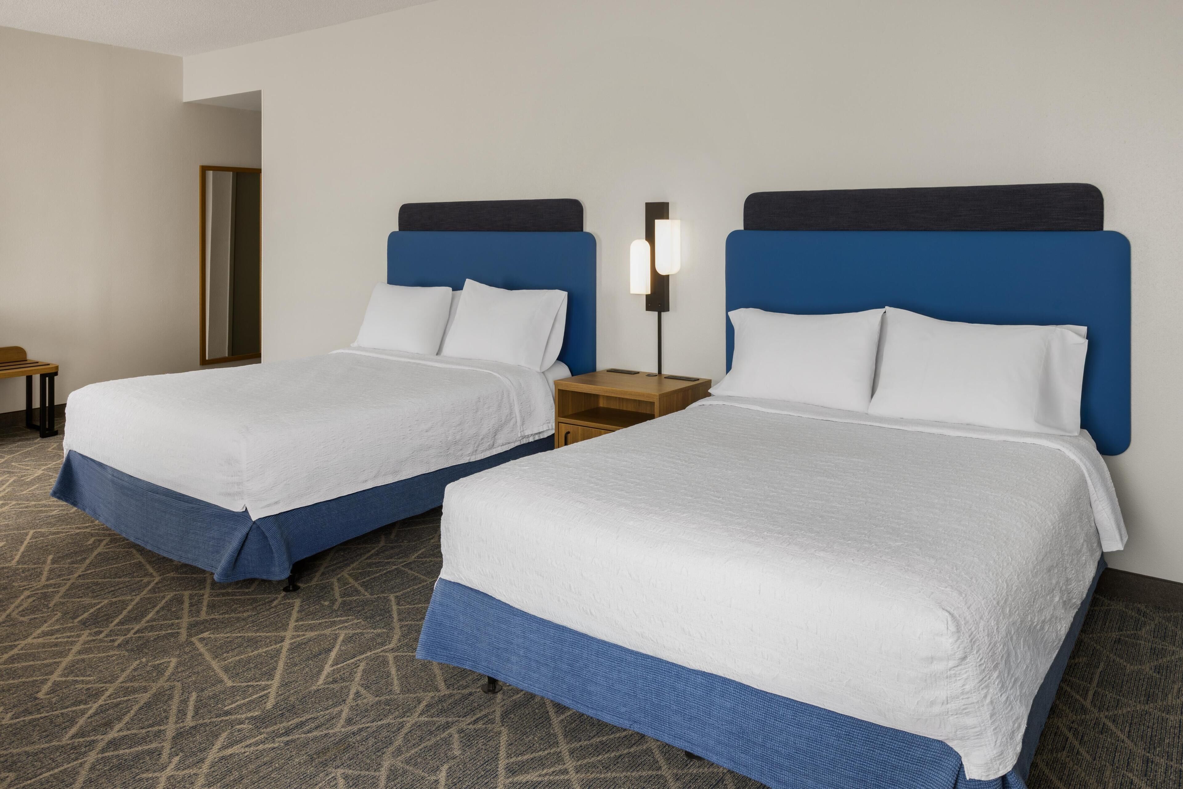 A hotel room with two beds, each with a blue headboard and white bedding. There is a nightstand with a lamp between the beds.