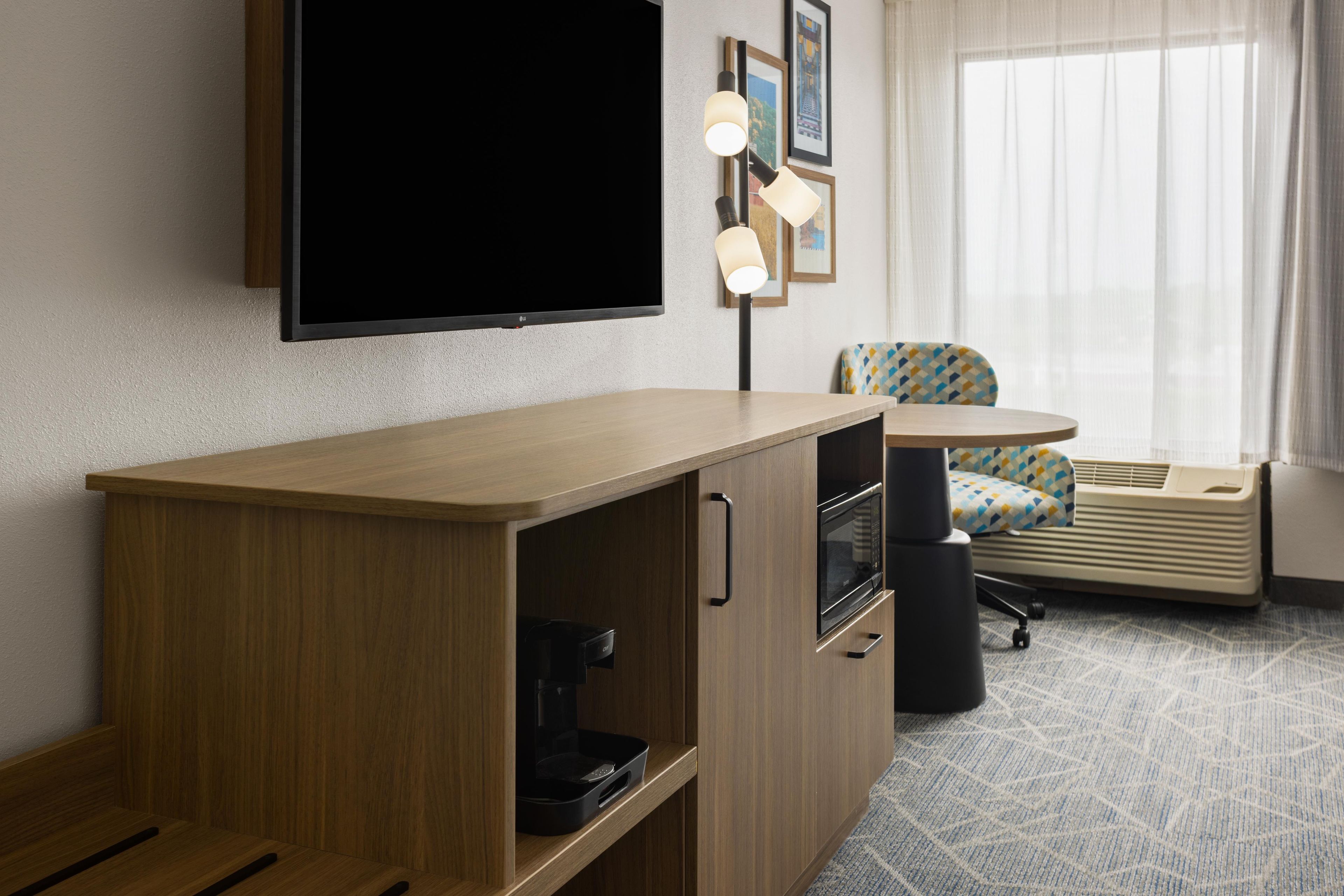 A hotel room with a wooden desk, a flat-screen TV, a coffee maker, a microwave, a chair, and a window with curtains.