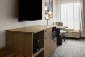 Habitación de hotel con escritorio de madera, televisor de pantalla plana, cafetera, microondas, una silla y ventana con cortinas.