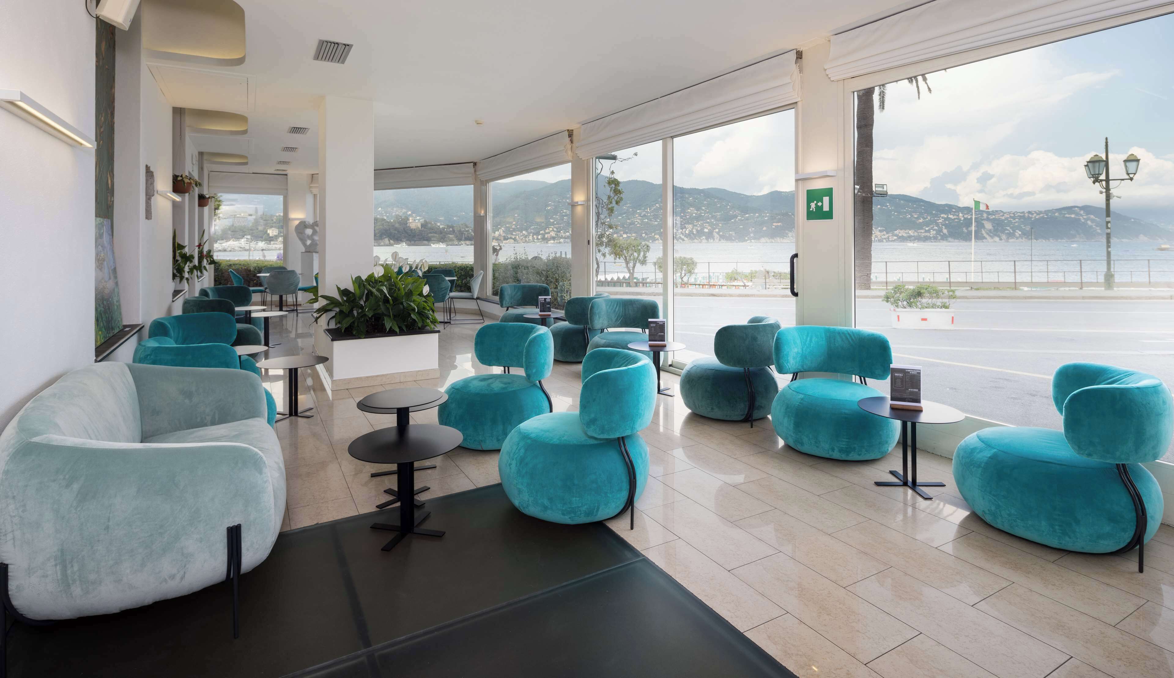 Um saguão de hotel moderno com assentos verde-água e branco, grandes janelas com vista para o mar e montanhas, e um design limpo e minimalista.