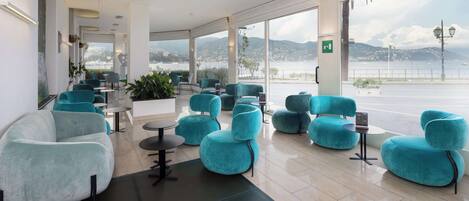 A modern hotel lobby with teal and white seating, large windows offering a view of the sea and mountains, and a clean, minimalist design.