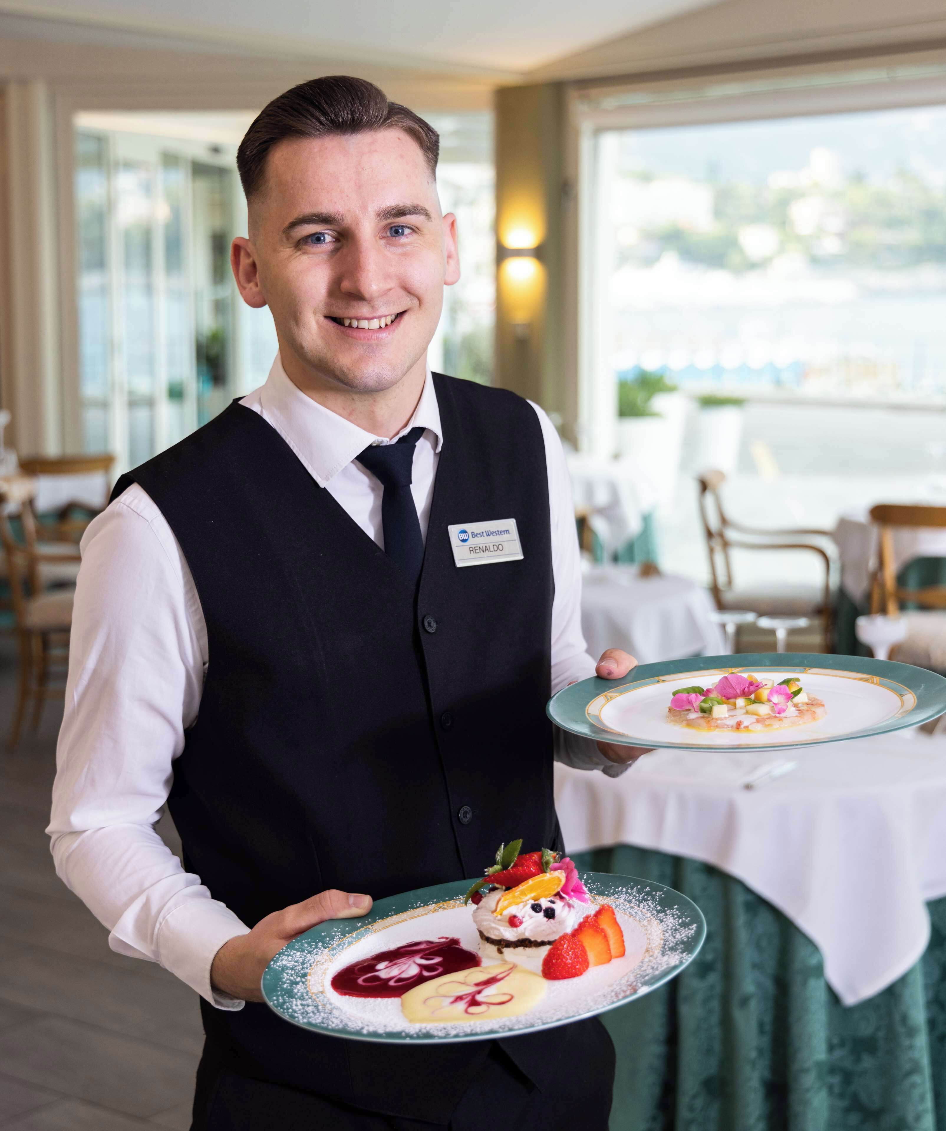 A waiter holding two plates of elegantly plated desserts in a well-lit dining area with a view of the outdoors.