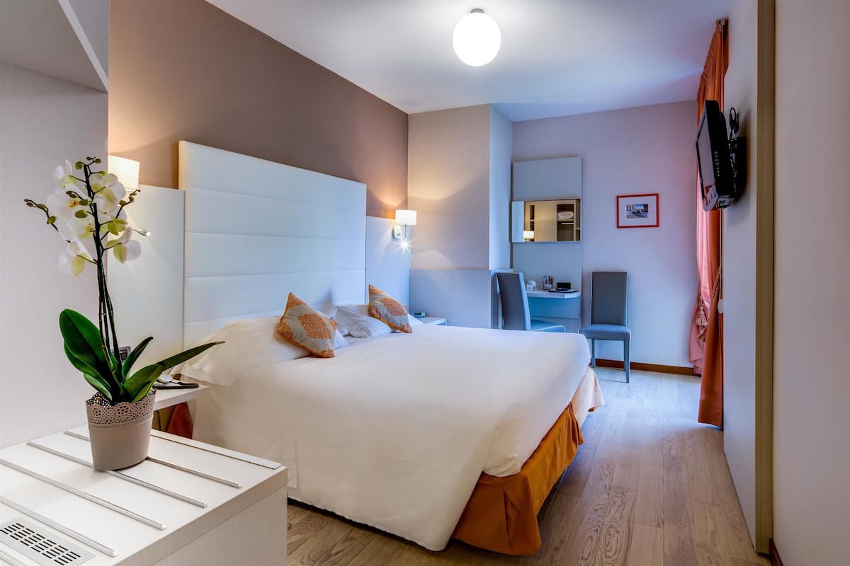 A hotel room with a large bed, a desk, a chair, a TV, and a potted plant.