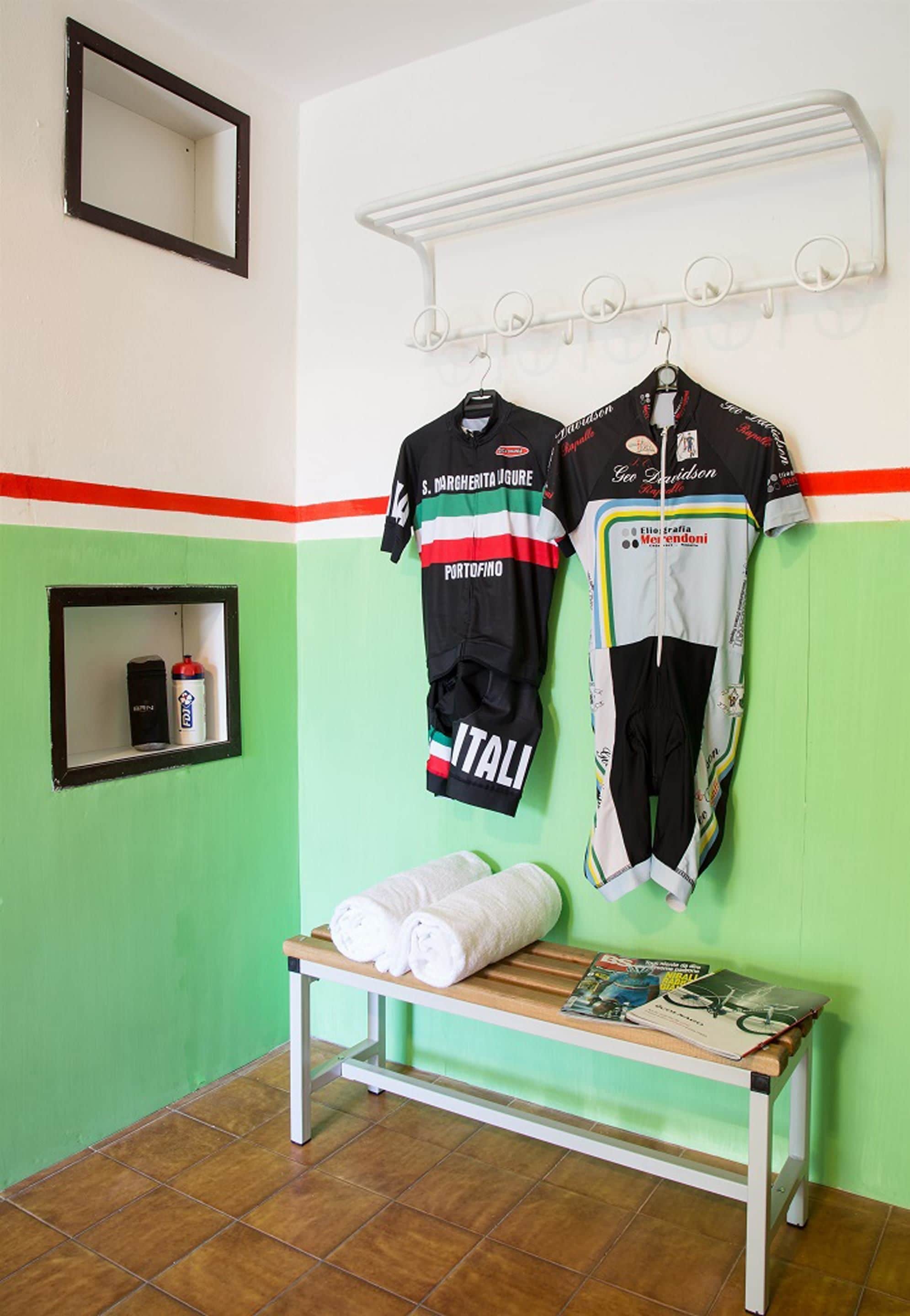 Una habitación con un banco, dos camisetas colgadas, un espejo y un estante con una botella de agua.