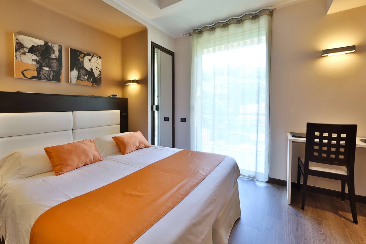 A hotel room with a large bed, a desk, and a chair. There are two paintings on the wall and a door with a mirror.