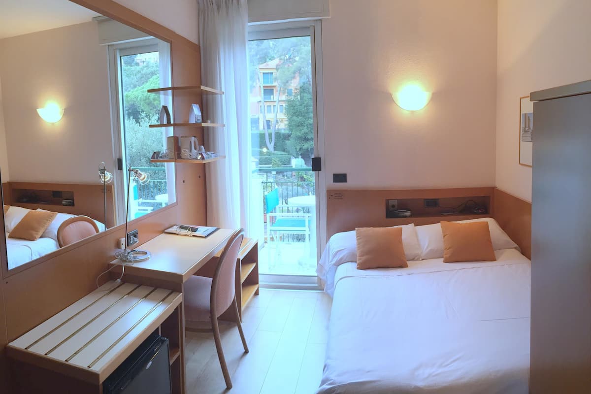 A hotel room with a bed, a desk, a chair, a mirror, and a balcony with a view of buildings and trees.