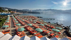 Une plage bordée de rangées de transats et de parasols, un rivage rocheux et une vue dégagée sur la mer et les montagnes lointaines.