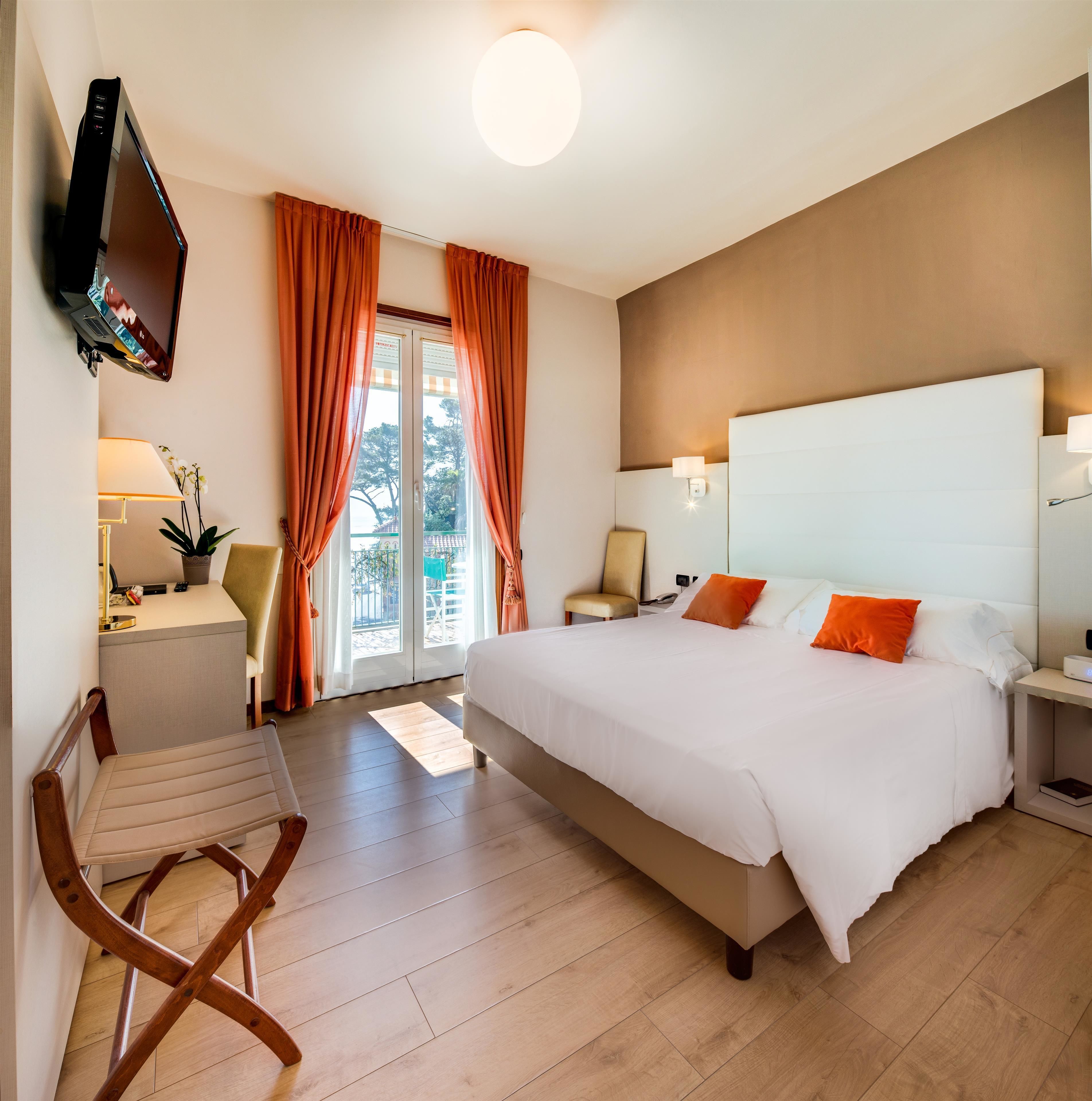 Una camera d'albergo con un letto grande, una scrivania con una lampada, una sedia e un televisore montato a parete.
