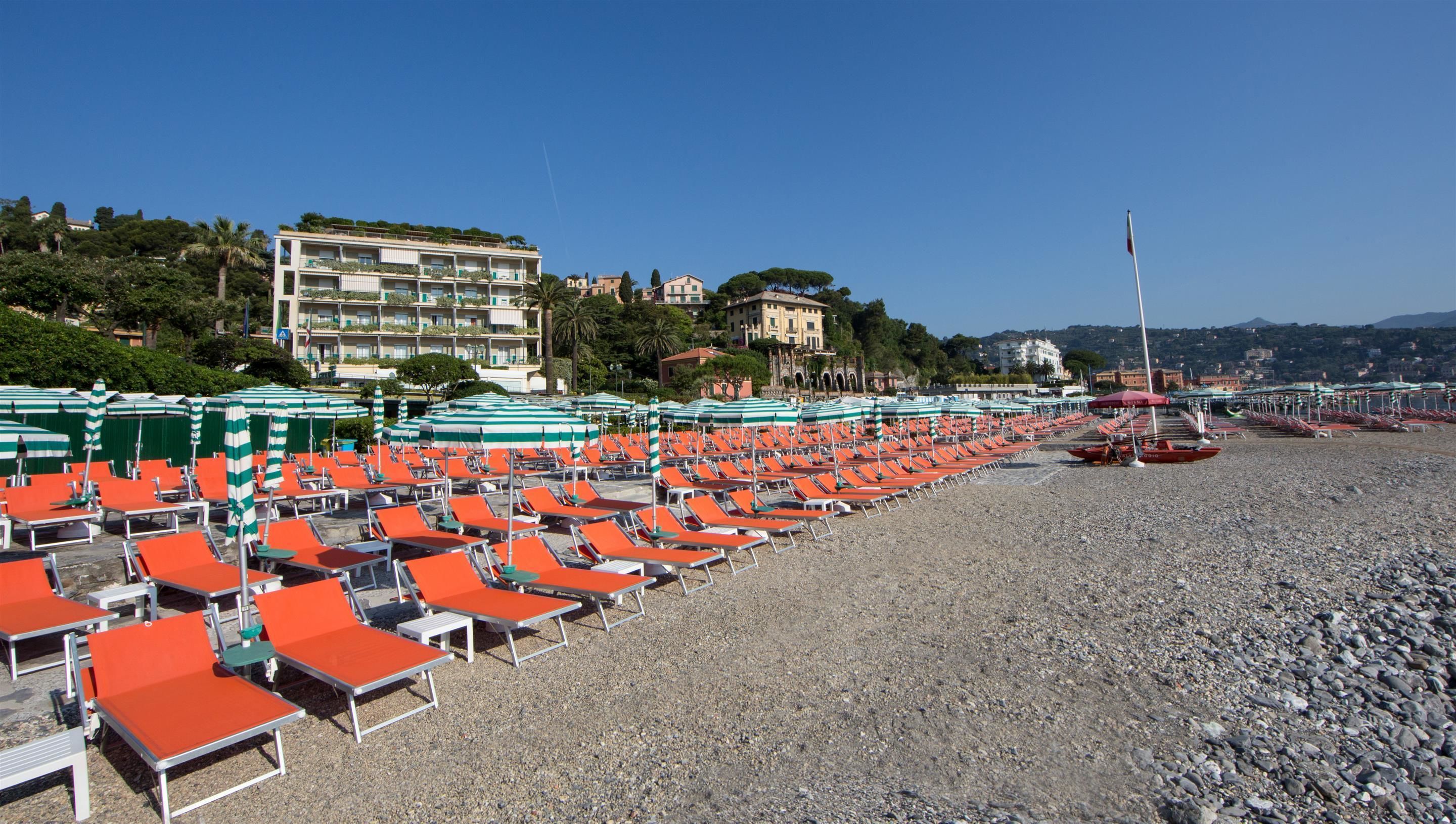 A beach with rows of orange lounge chairs and green umbrellas, a rocky shoreline, and buildings in the background.