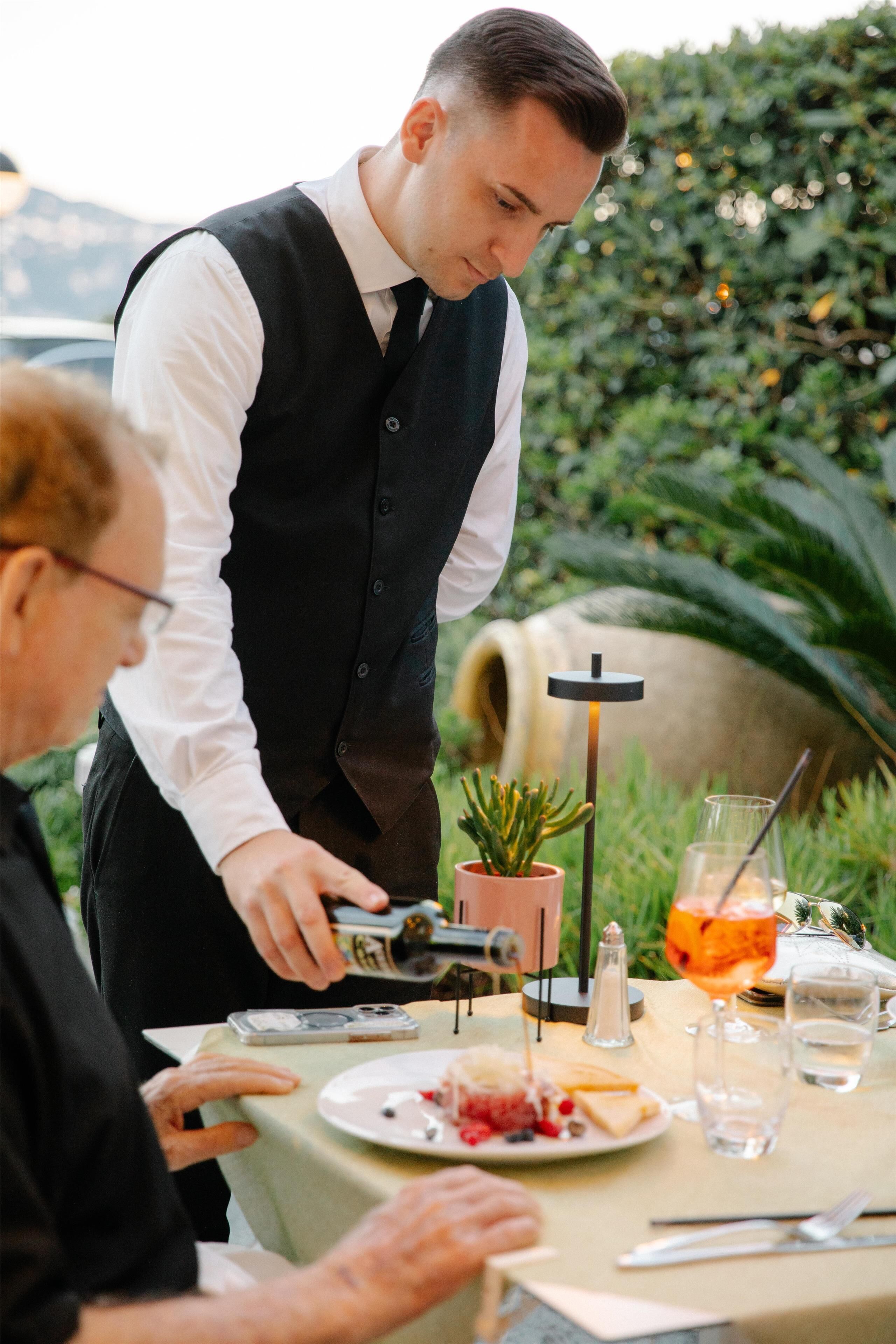 Un serveur apporte des plats à une table en plein air, avec en arrière-plan un paysage verdoyant et des montagnes.