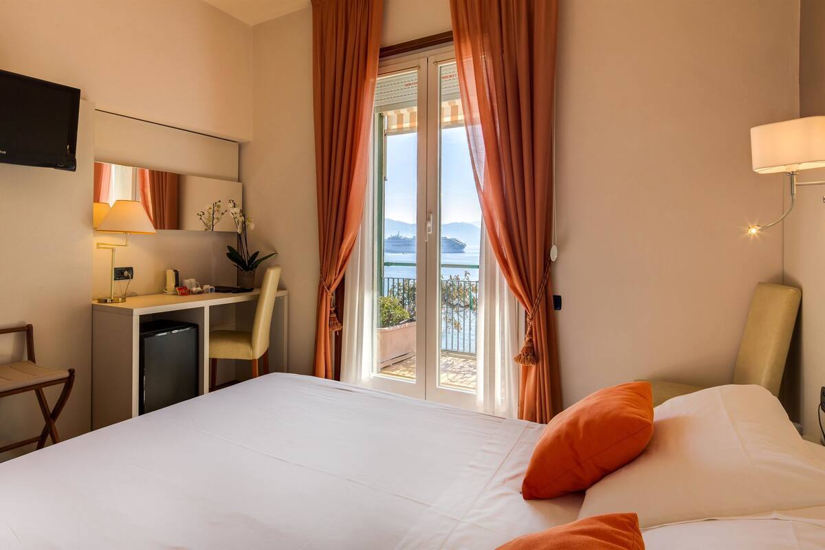 A hotel room with a bed, desk, chair, TV, and a view of the sea through the window.