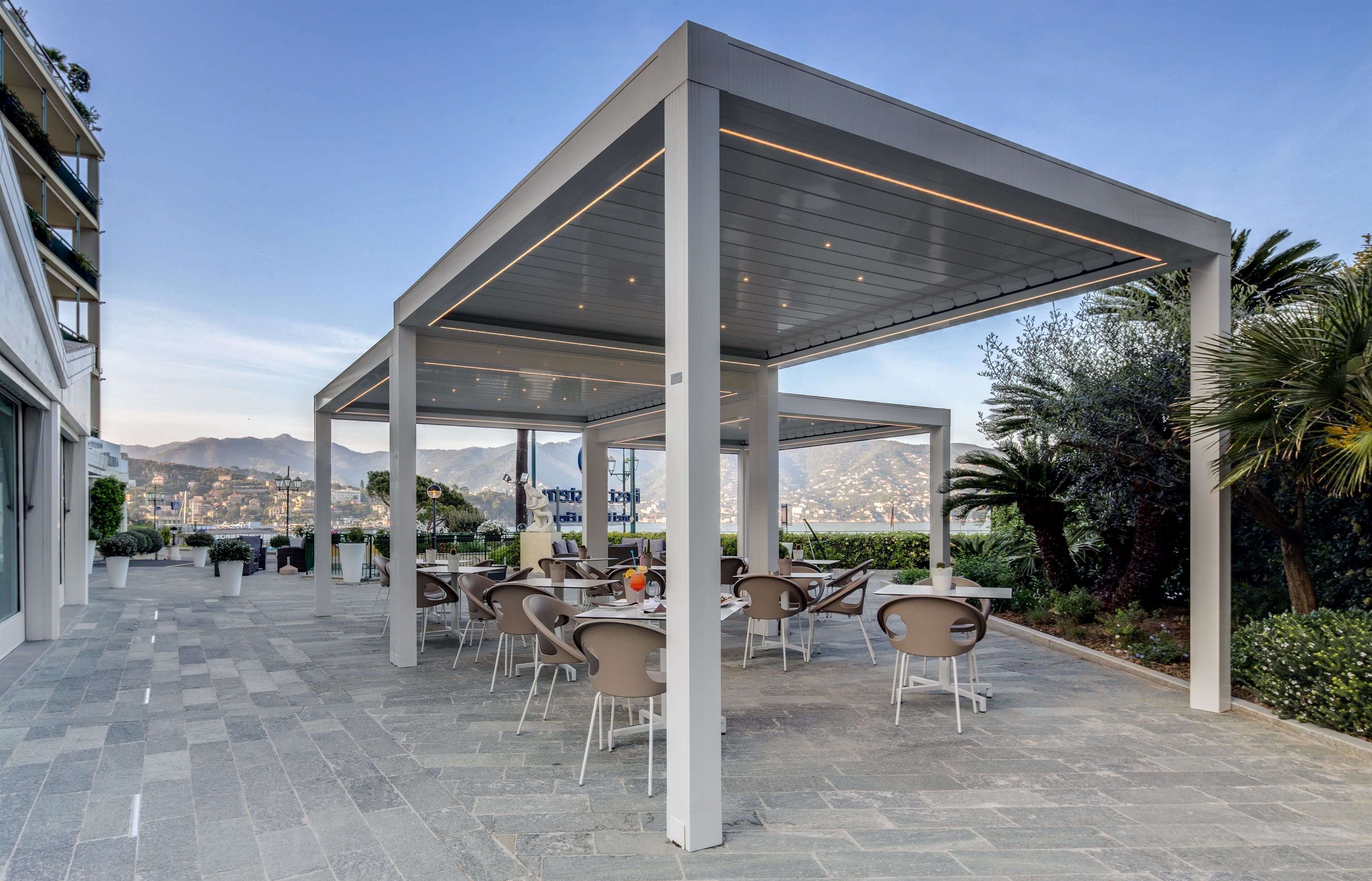 An outdoor patio with tables and chairs, a covered area with a roof, and a view of mountains in the background.