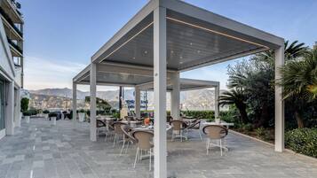An outdoor patio with tables and chairs, a covered area with a roof, and a view of mountains in the background.