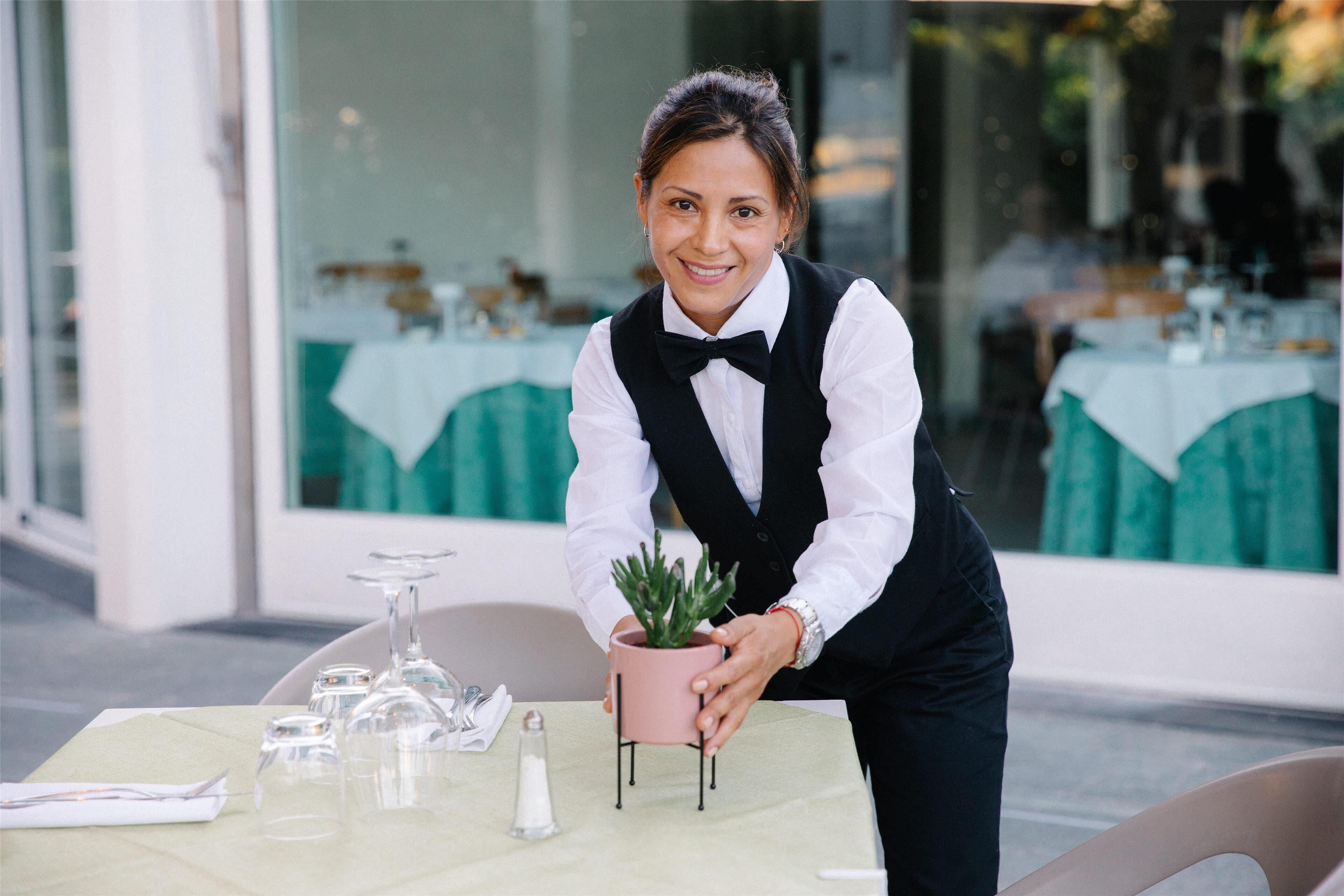 Un serveur, vêtu d’un gilet noir et d’une chemise blanche, tient un pot de fleurs sur une table recouverte d’une nappe verte, où se trouvent des verres transparents et un salière.