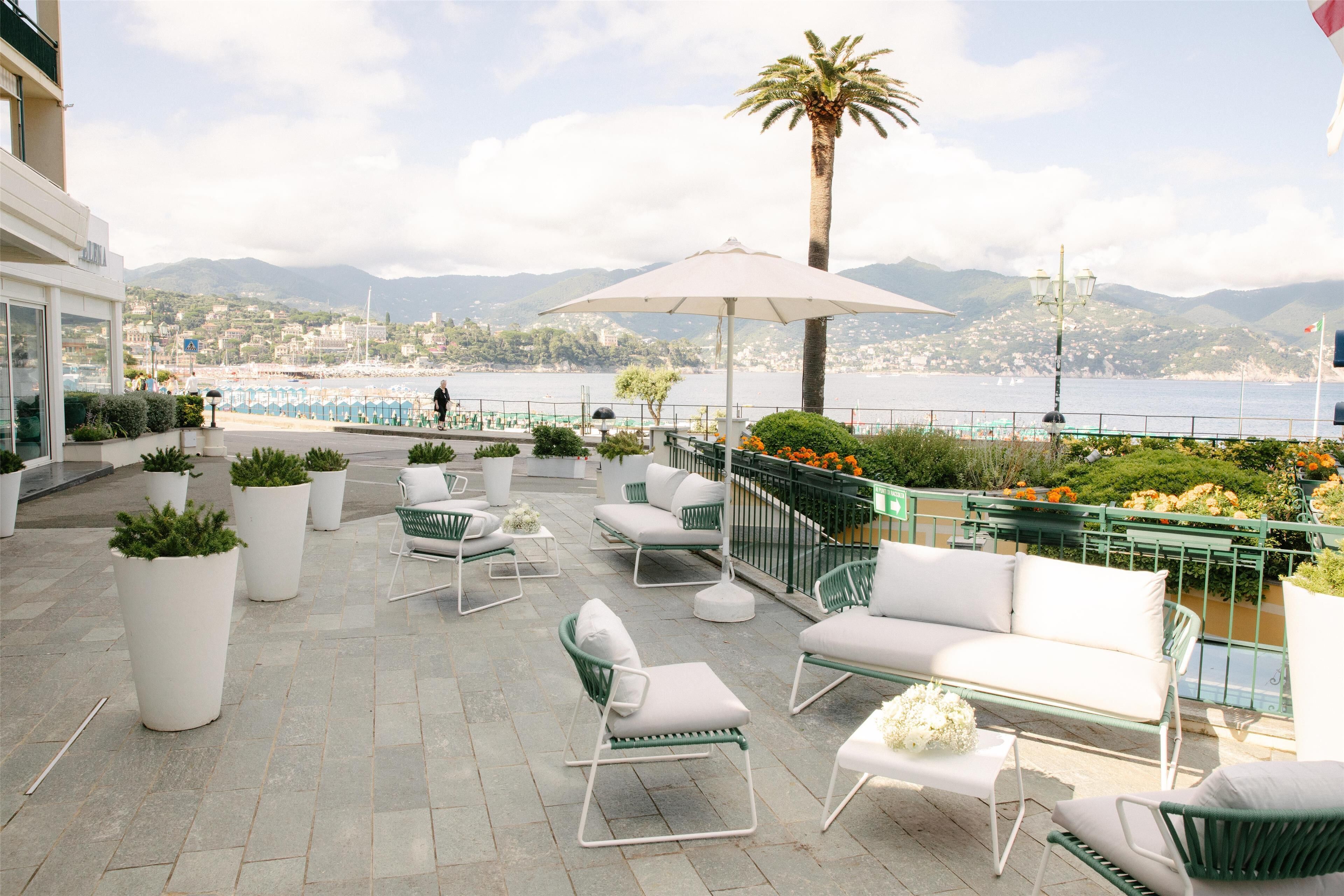 An outdoor seating area with white furniture, potted plants, and a view of the sea and mountains.