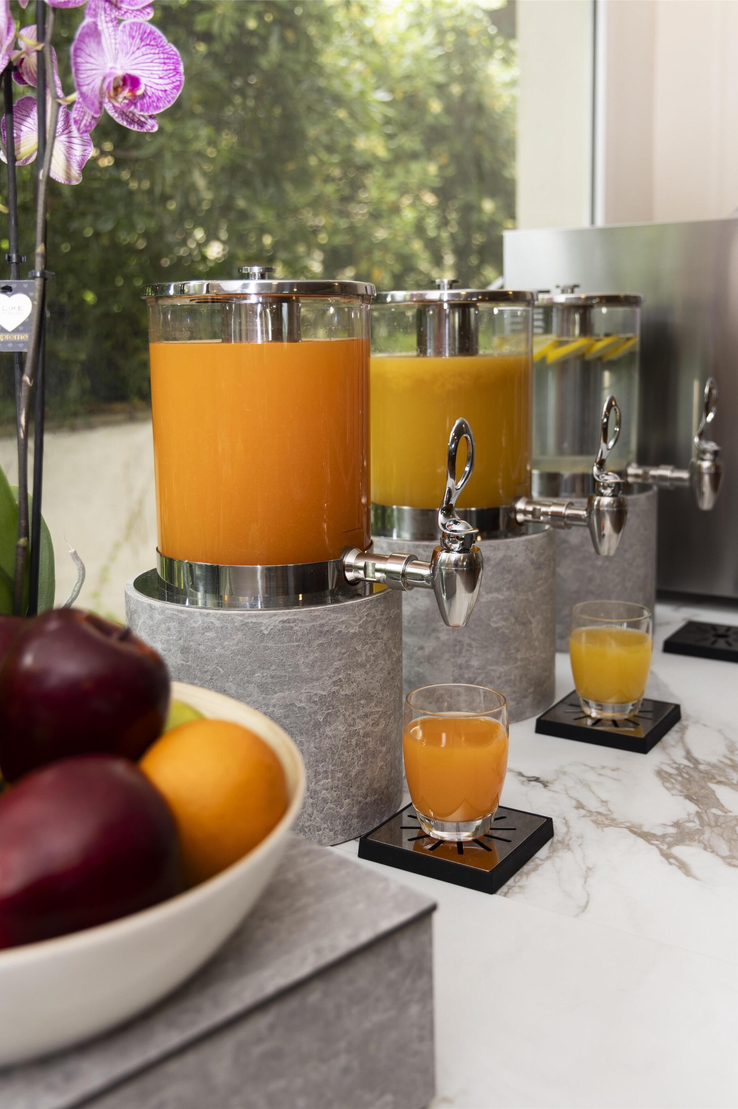 A juice bar with multiple dispensers, a bowl of fruit, and a glass of juice on a marble countertop.