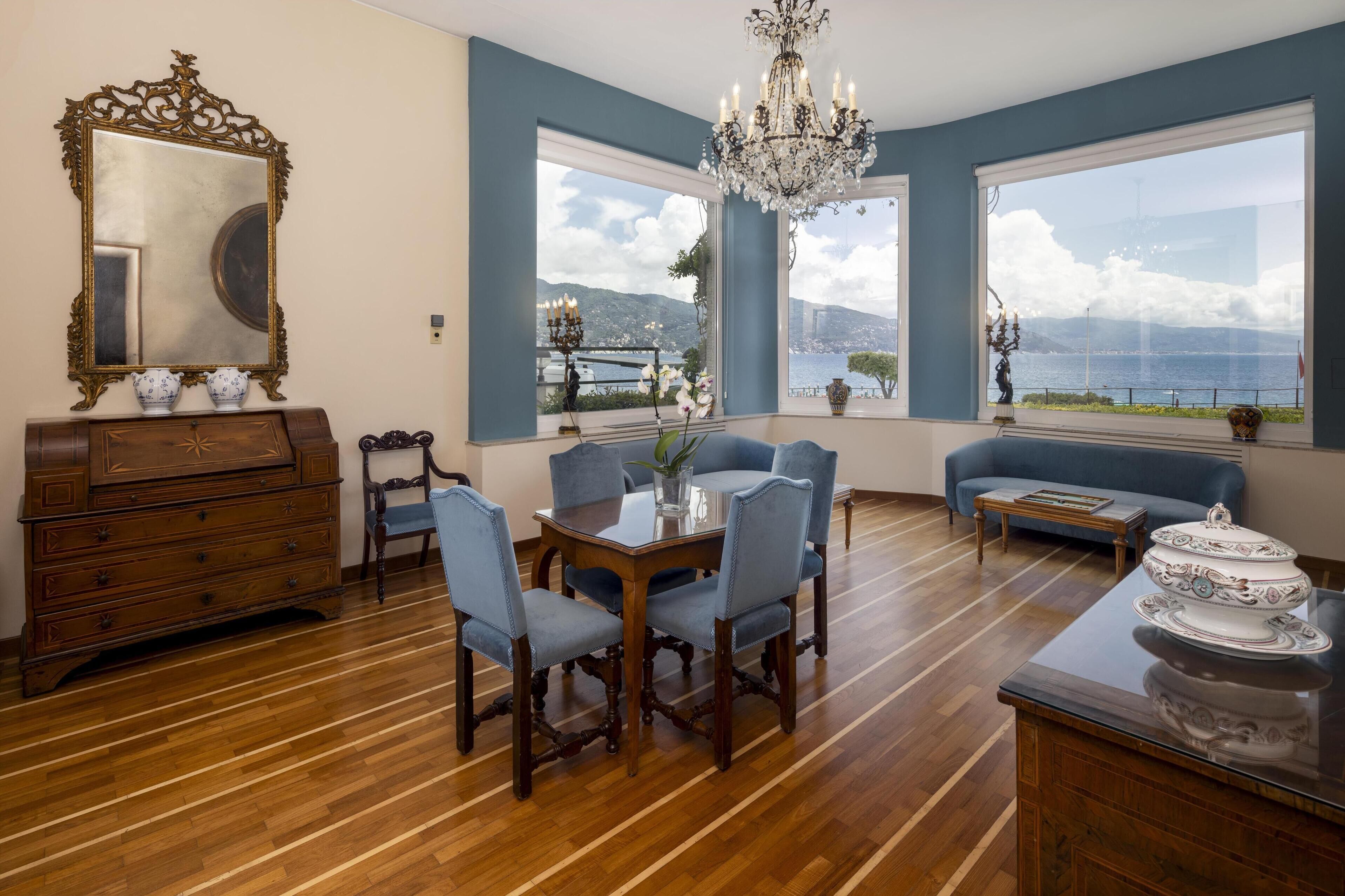 A dining area with a wooden table and chairs, a chandelier, and large windows with a view of the sea and mountains.