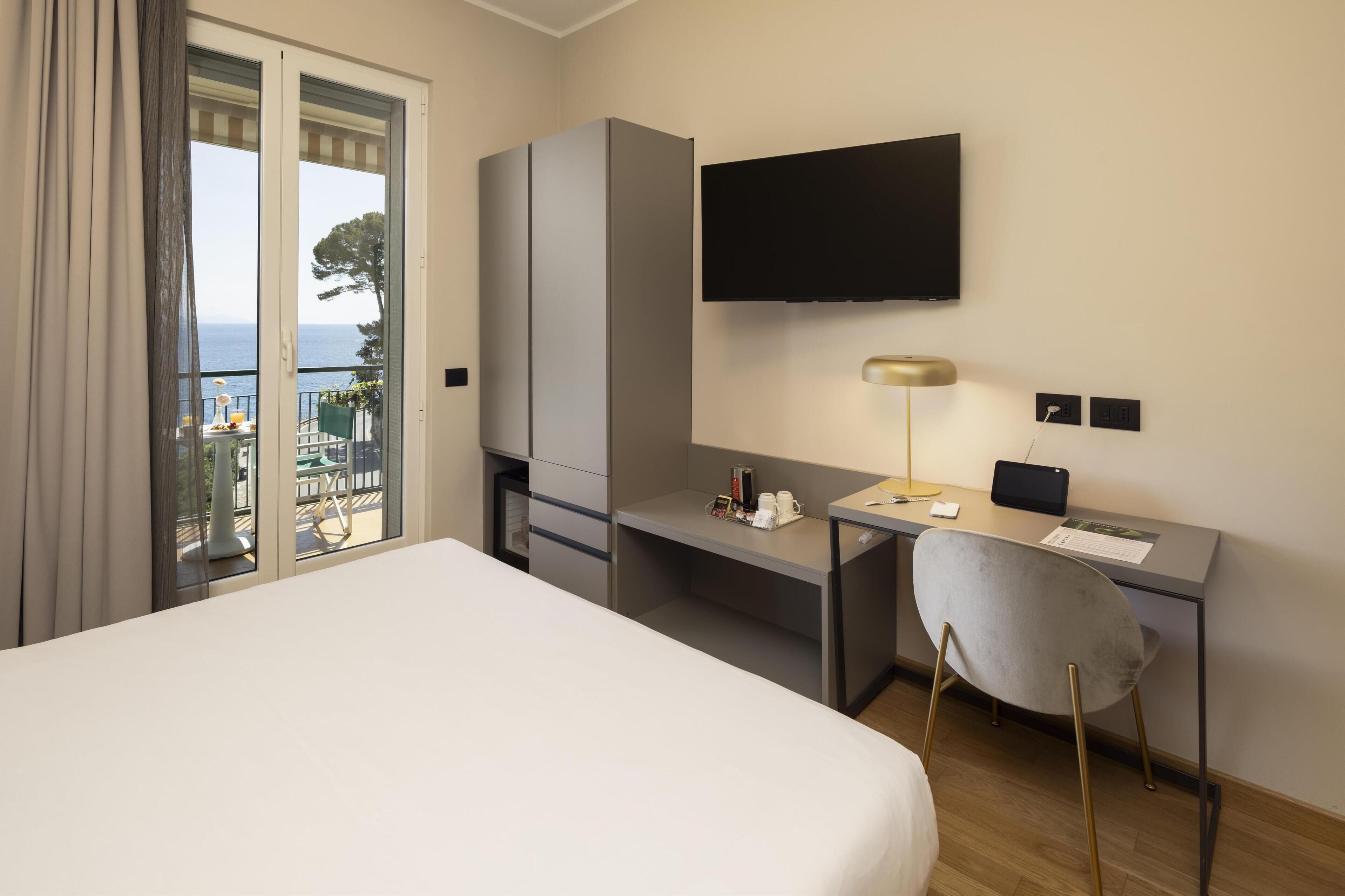 Habitación de hotel con cama, escritorio, silla, televisor y balcón con vista al mar.