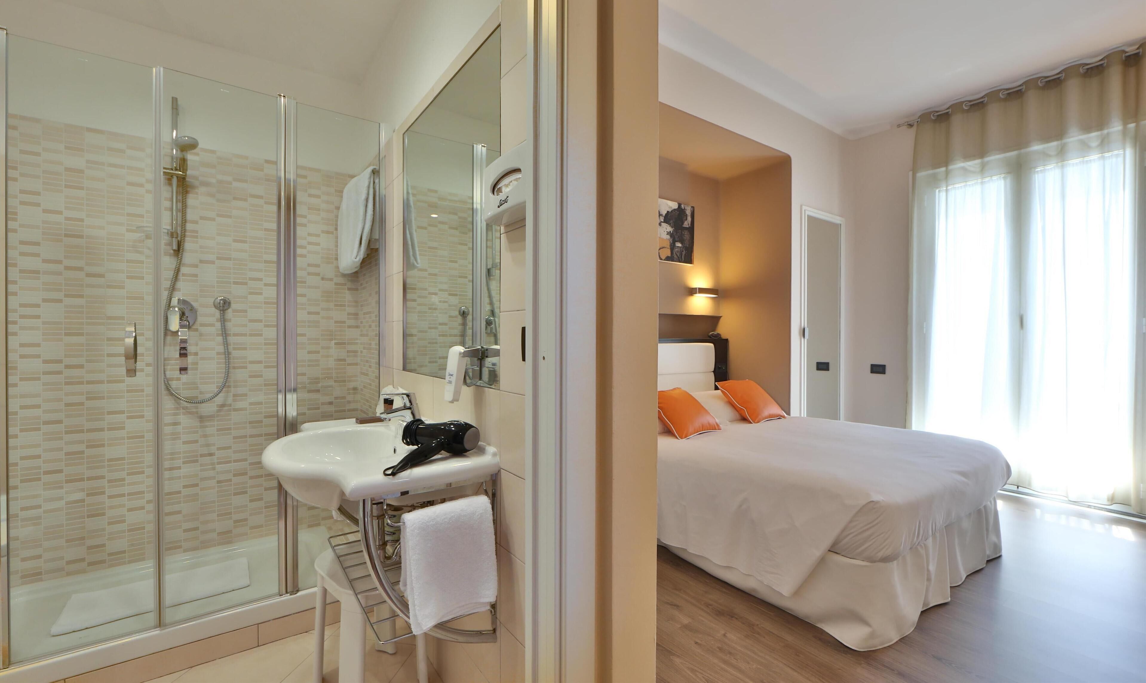 A hotel room with a glass-enclosed shower, a sink, a bed with orange pillows, and a large window with curtains.