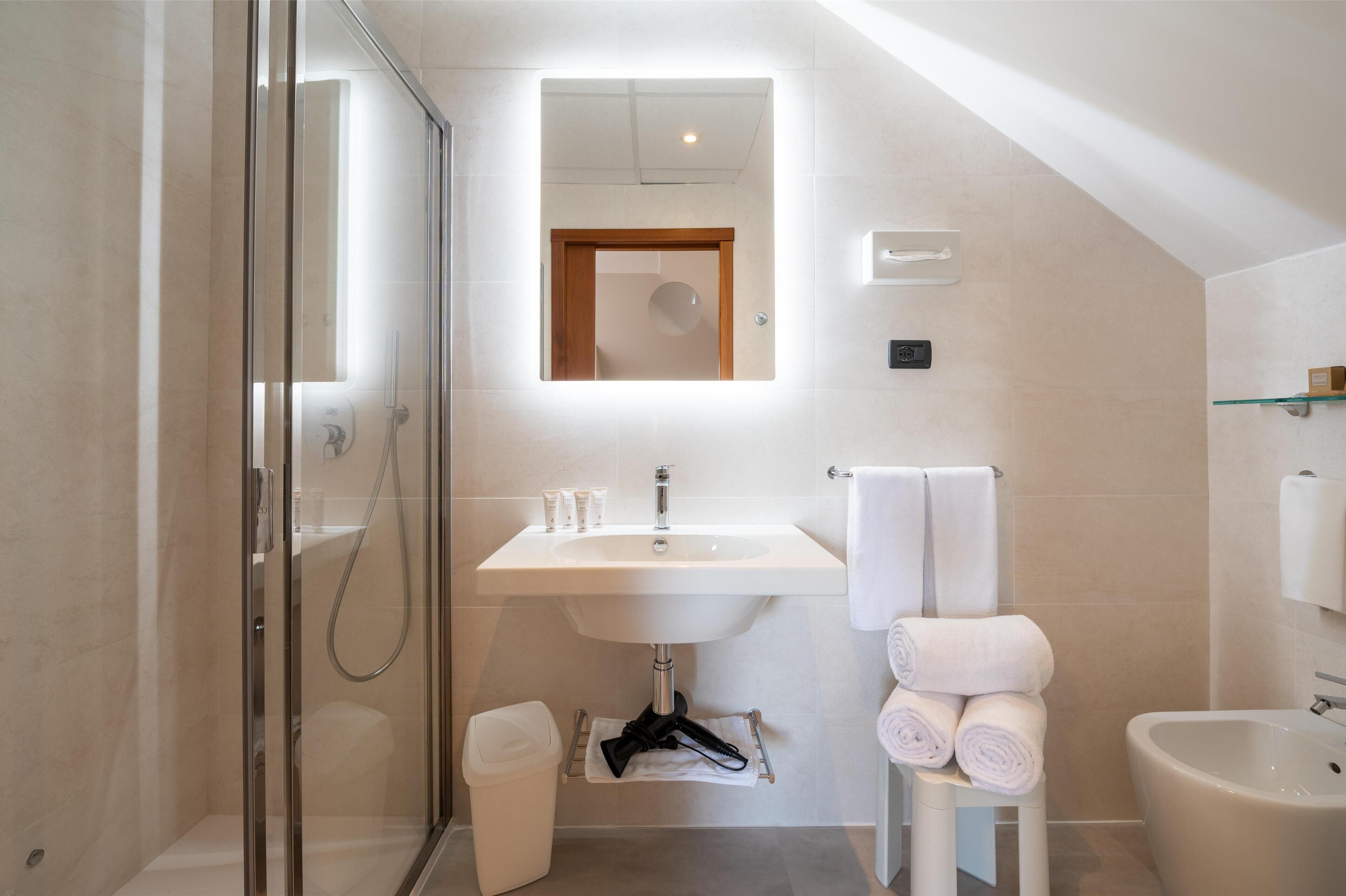 Une salle de bain moderne avec une douche à parois de verre, un lavabo avec un miroir au-dessus et un bidet. Des serviettes sont pliées et posées sur une petite table à côté du lavabo.