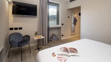 Ein Hotelzimmer mit Bett, Wandfernseher, Stuhl, kleinem Tisch mit Pflanze und Blick ins Freie durch ein Fenster.
