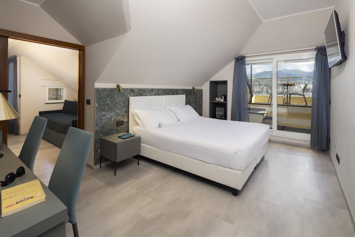 A bedroom with a large bed, a desk with a chair, a nightstand, and a view of the city through a balcony.