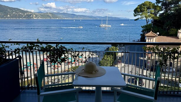 Un balcon avec une table et des chaises, donnant sur une zone côtière où l’on peut apercevoir des bateaux et, au loin, une chaîne de montagnes.