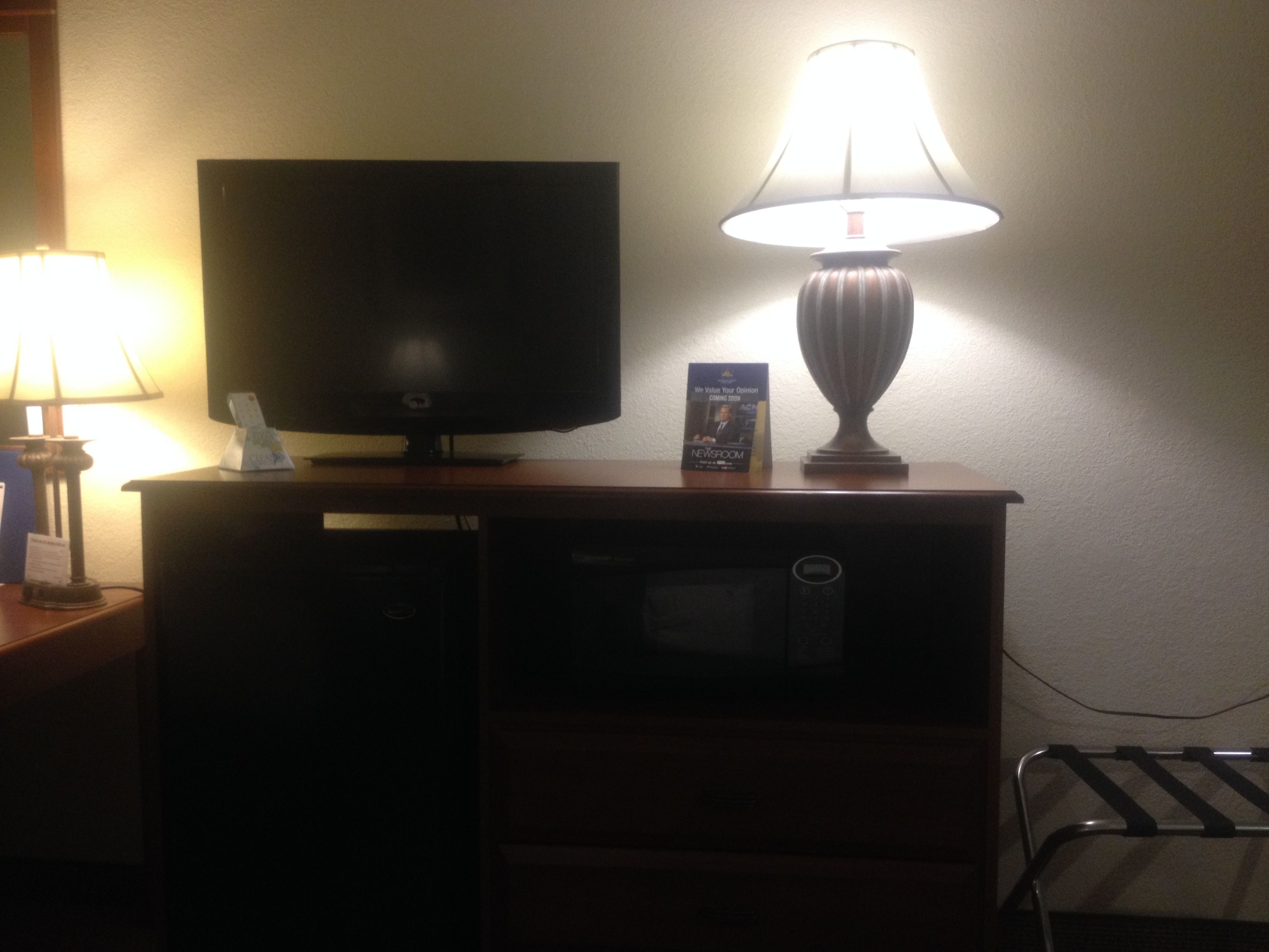 Una habitación de hotel con un televisor sobre un mueble de madera, una lámpara en una mesita auxiliar y una pequeña señal en el mueble.