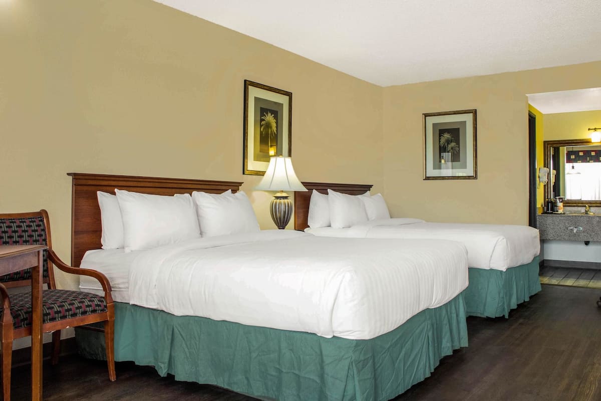 A hotel room with two beds, a desk, a chair, and a nightstand with a lamp.