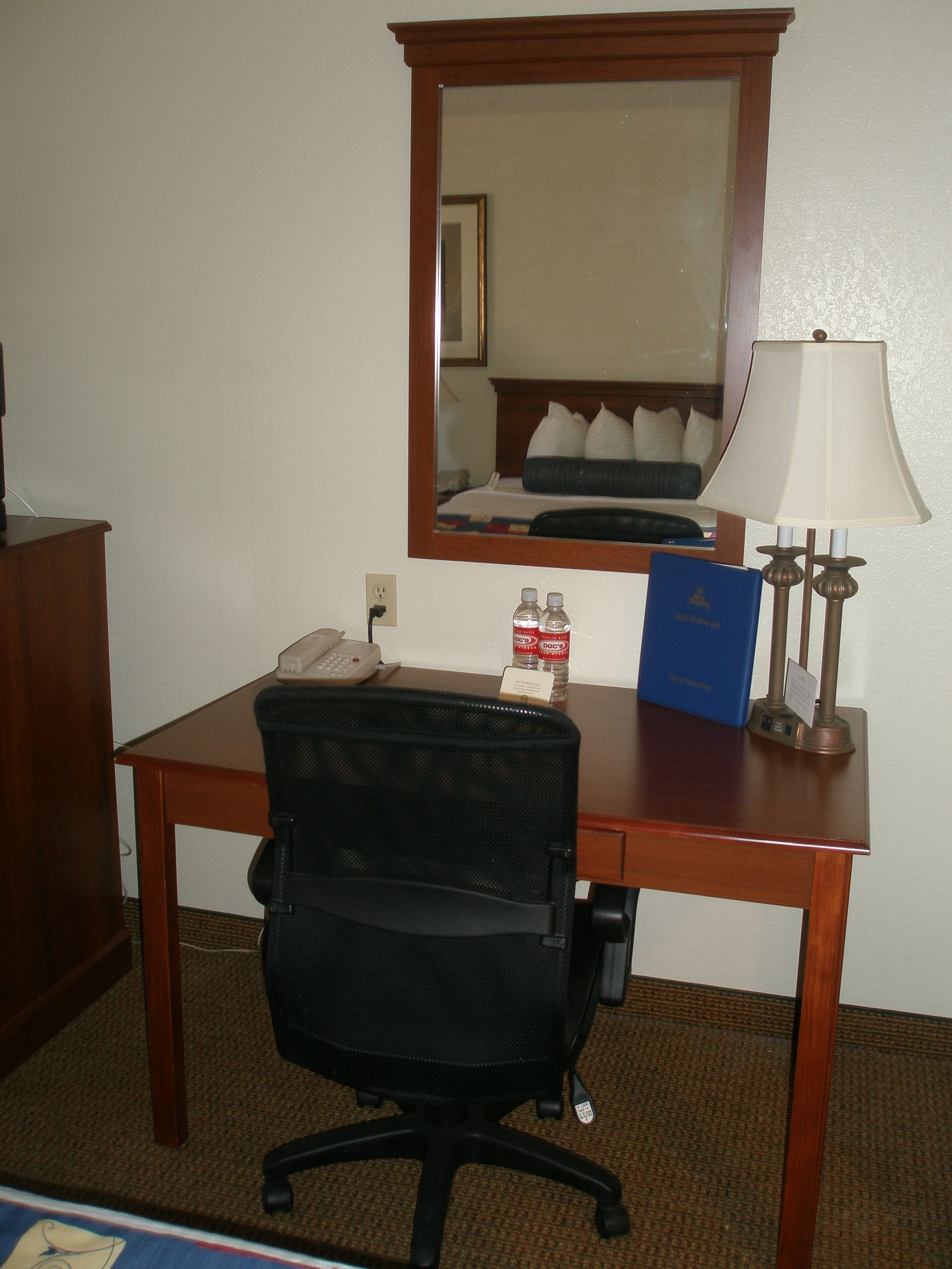 Une chambre d’hôtel avec un bureau en bois, une chaise de bureau noire, une lampe, un miroir, un téléphone et un livre bleu posé sur le bureau.