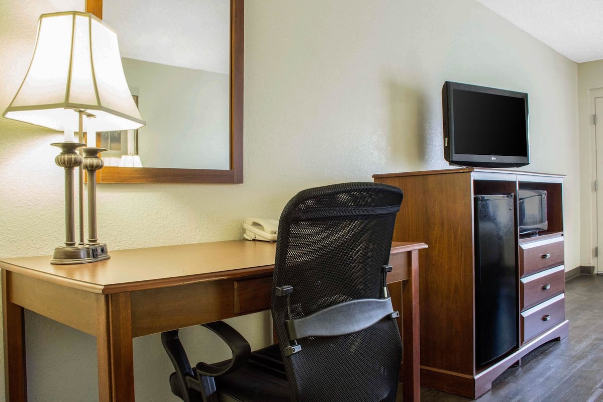 A hotel room with a wooden desk, a black mesh office chair, a television on a wooden cabinet, and a lamp on the desk.