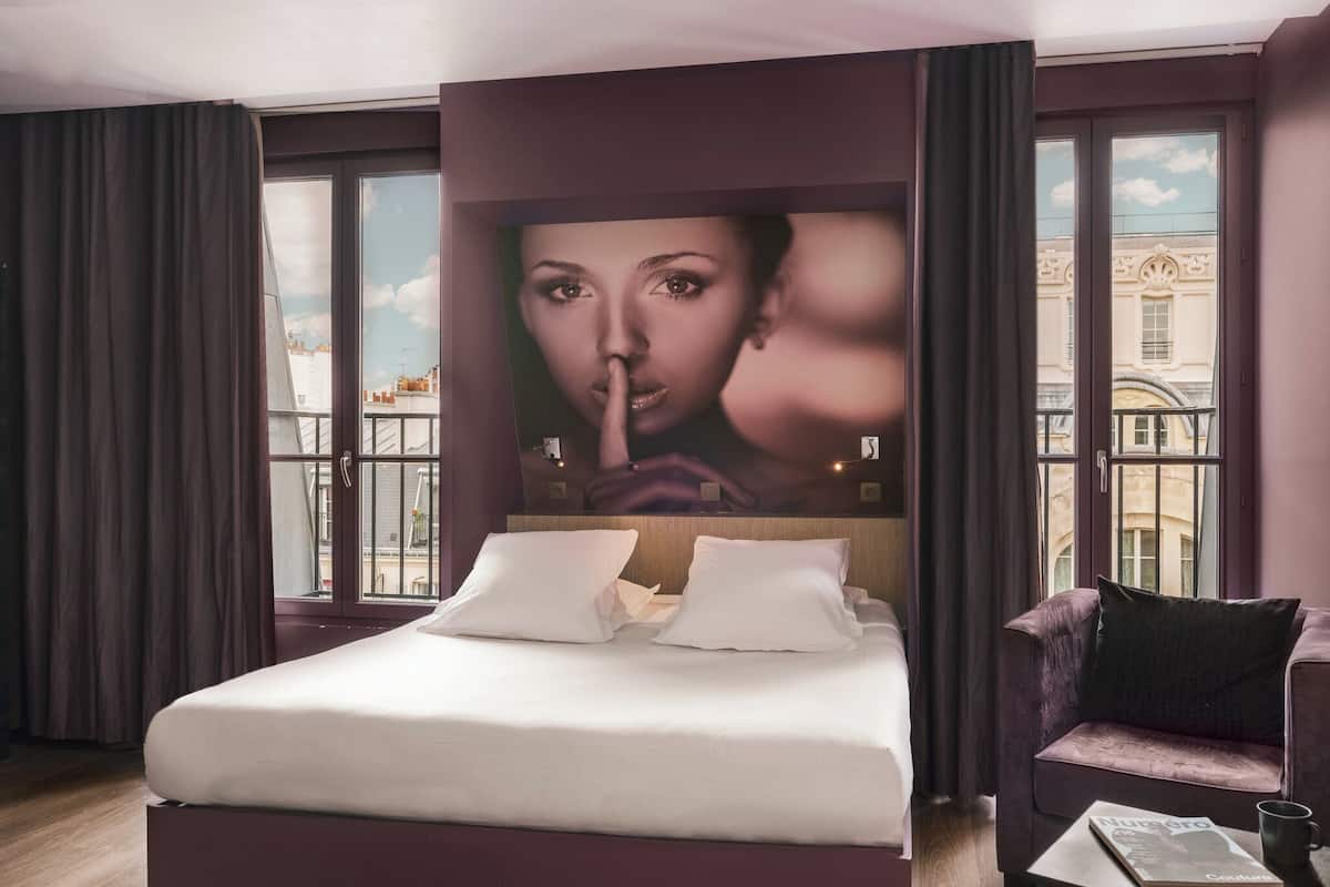Een moderne hotelkamer met een groot bed, een bank en uitzicht op de stad door grote ramen.