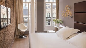 Een hotelkamer met een bed, een stoel, een klein tafeltje met een bloevas en uitzicht op gebouwen door grote ramen.
