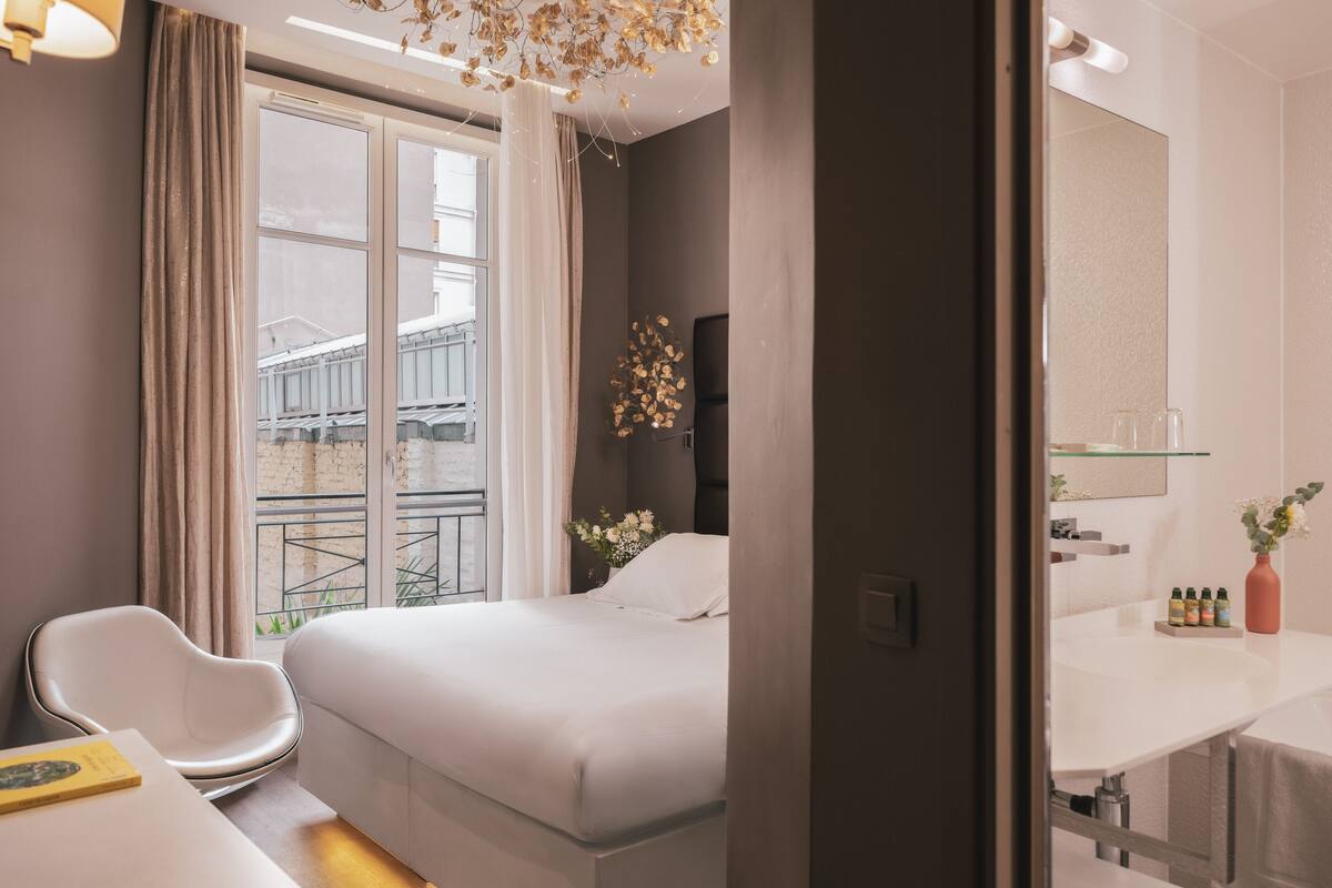 Een moderne hotelkamer met een groot bed, een bureau met stoel, een balkon met uitzicht op gebouwen en een badkamer met spiegel en toiletartikelen.