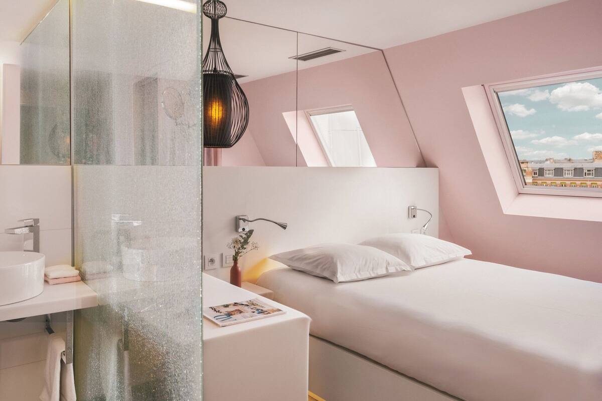 Een moderne slaapkamer met een bed, een nachtkastje met lamp, een raam met uitzicht op gebouwen, en een badkamer met wastafel en spiegel.