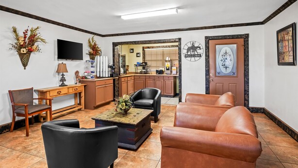 Un salon avec une table basse, un canapé en cuir et un téléviseur. Au fond, on aperçoit une cuisine avec des placards et une porte.