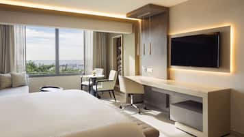 A hotel room with a large bed, a desk, a chair, a TV, and a view of the city.