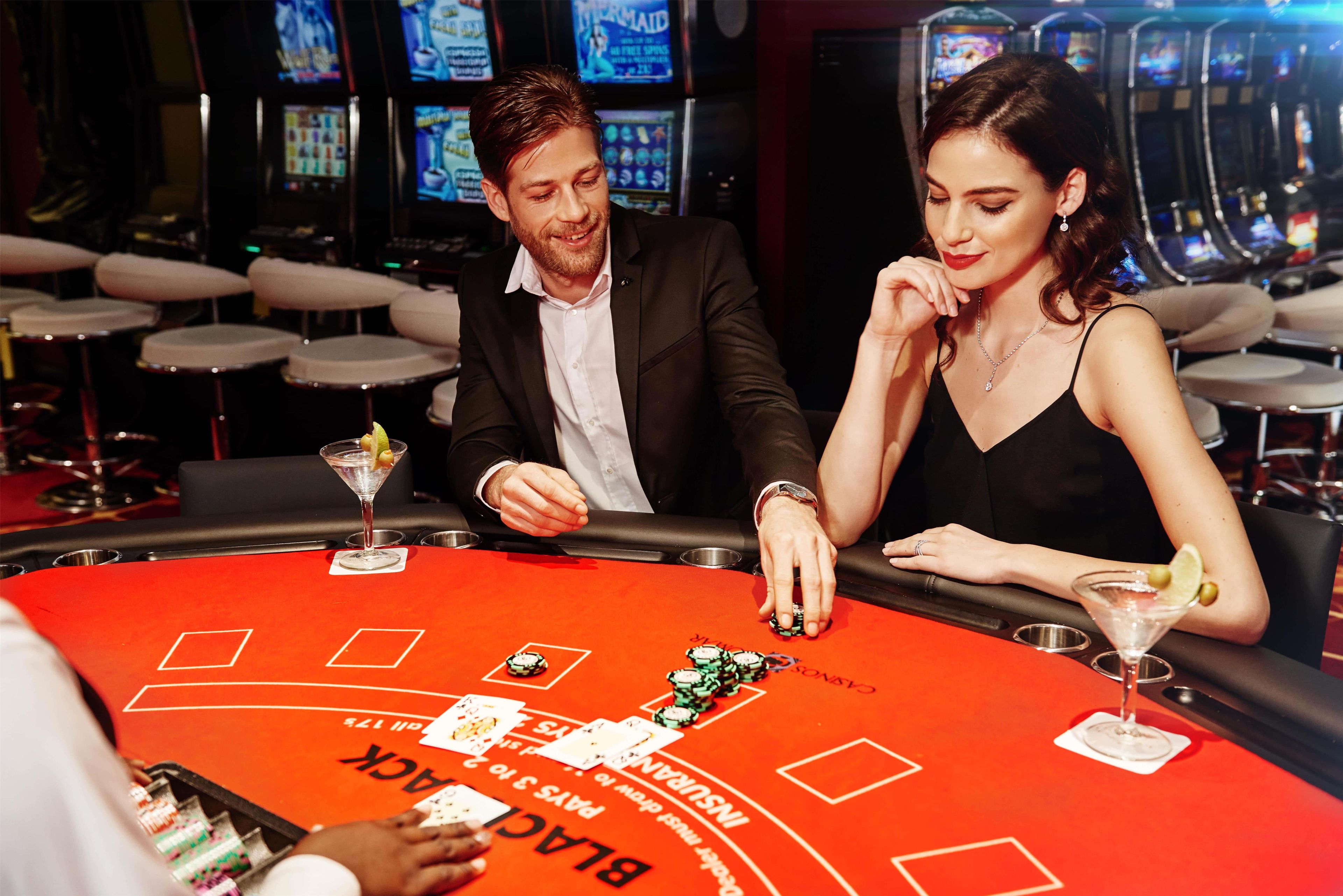 A casino blackjack table with chips and cards, a woman and a man playing, and slot machines in the background.