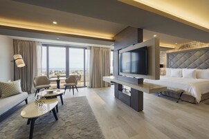 A modern hotel room with a large TV, a sofa, a dining table, and a bed.