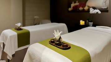 Un cuarto de spa con dos camillas, cada una con un mantel blanco y una corrida verde. Hay velas y una bandeja pequeña sobre las camillas.