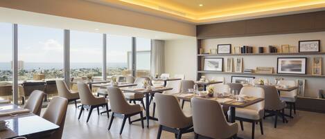A modern dining area with tables and chairs, large windows offering a city view, and a wall with framed artwork.