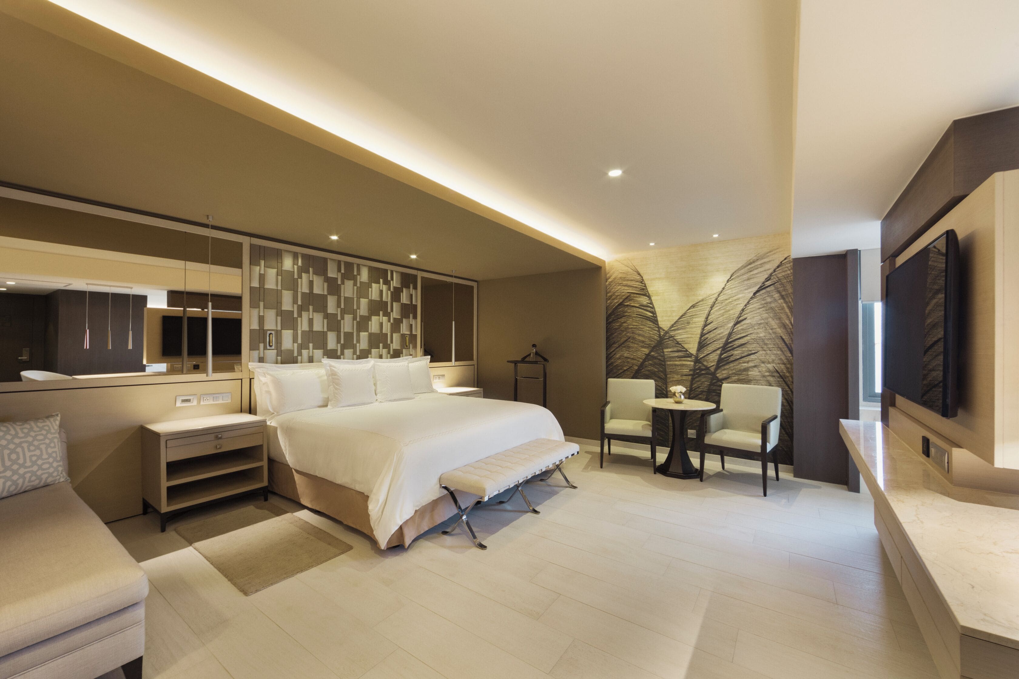 Une chambre d’hôtel moderne dotée d’un grand lit, de tables de chevet, d’un coin salon avec des fauteuils, d’une petite table et d’une grande œuvre d’art accrochée au mur.