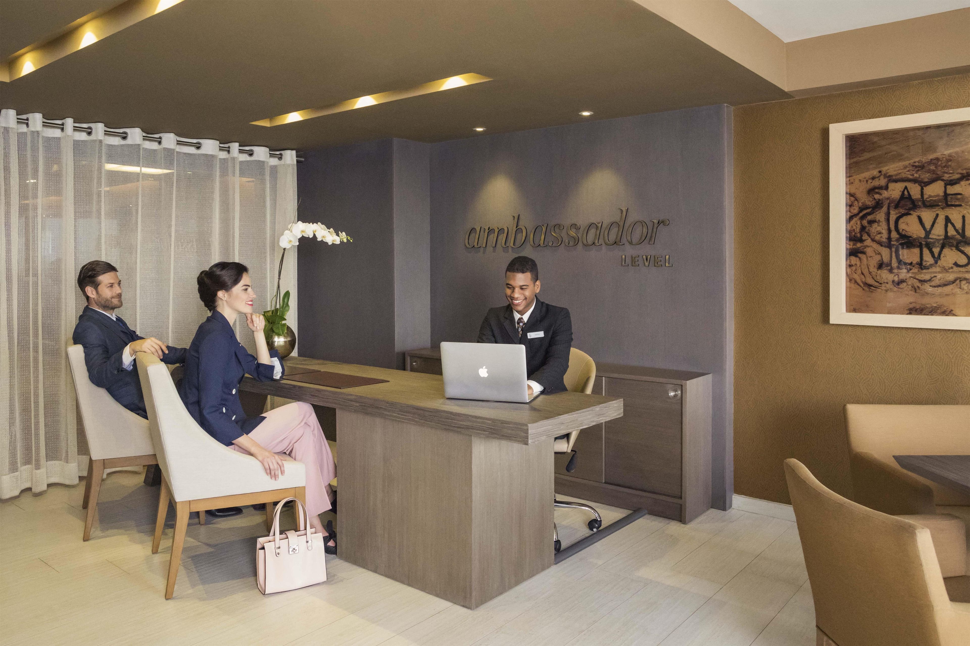 A reception desk with a nameplate reading 'Ambassador' and 'Levell'. There are three people seated on either side of the desk.