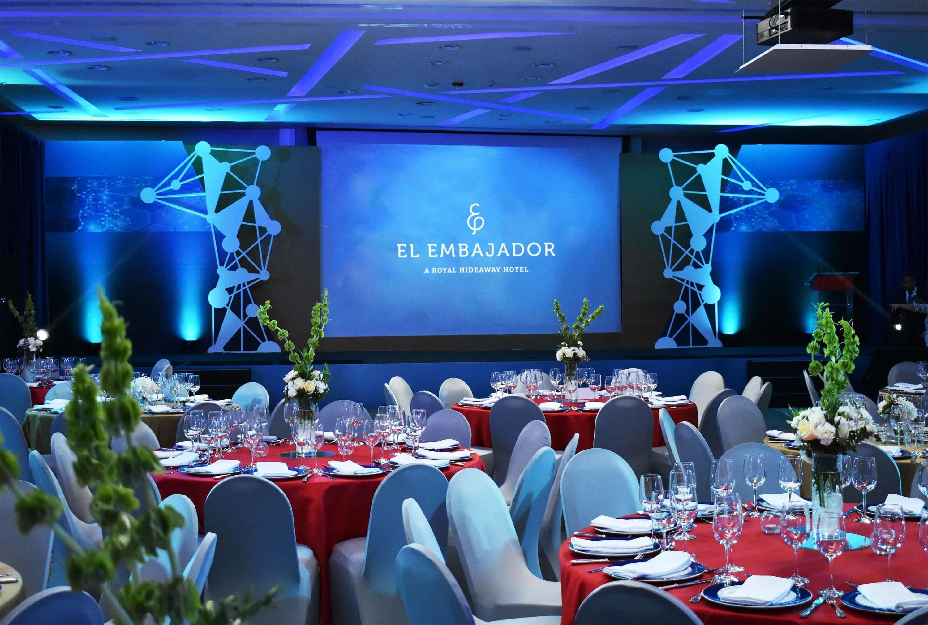 Un salón de banquetes en el hotel El Embajador, con mesas dispuestas para un evento formal, una pantalla grande mostrando el nombre del hotel y una moderna lámpara con diseño geométrico.