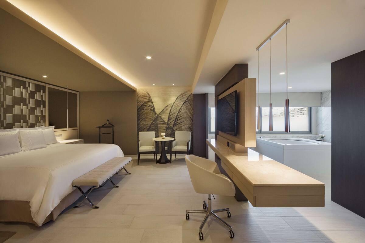 Ein modernes Hotelzimmer mit einem großen Bett, einem Schreibtisch mit Stuhl, einem kleinen Tisch, einem Fernseher und einer Badewanne.