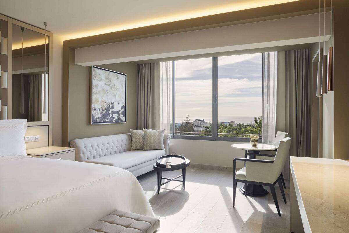 Ein Hotelzimmer mit einem großen Bett, einer Couch, einem runden Tisch und Blick auf die Stadt durch große Fenster.