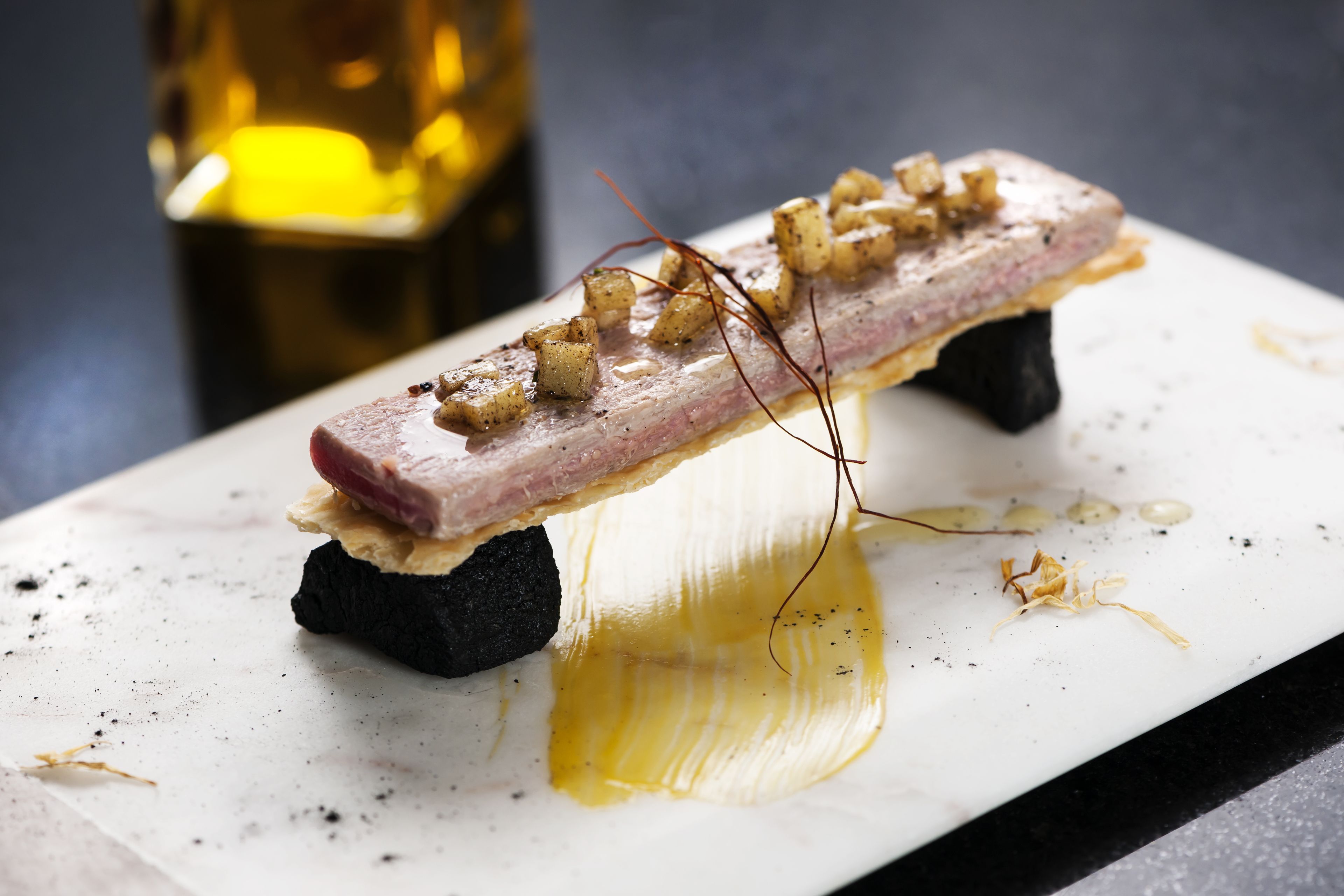 A dish with a slice of meat, diced potatoes, and a garnish on a white plate with a black base.