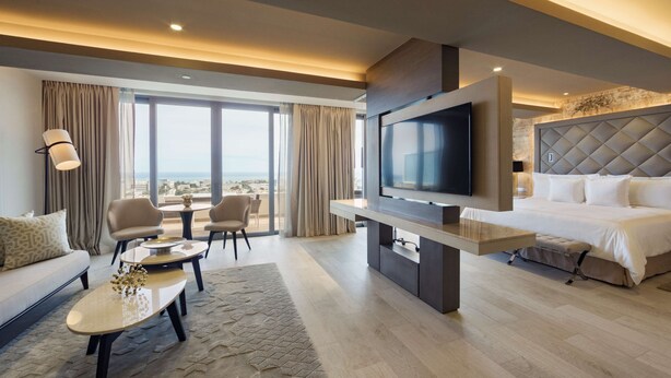 A modern hotel room with a large bed, a flat-screen TV, a sofa, a coffee table, and a view of the city.