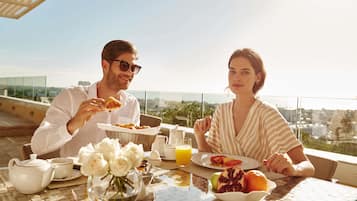 Un couple est assis à une table avec vue sur la ville, devant un assortiment de plats pour le petit-déjeuner et un vase rempli de fleurs.